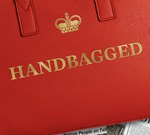 Handbagged is embossed in gold on a red handbag. The handbag is in front of black and white newspaper cuttings.