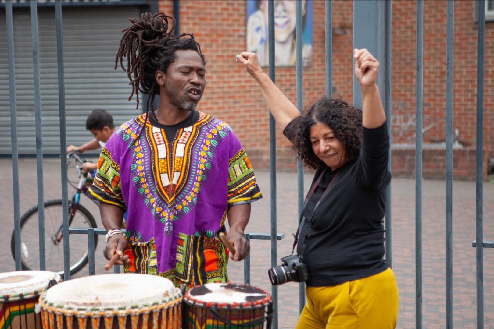 A man in a colourful patterned dashiki plays drums outdoors while a smiling woman with a camera around her neck dances beside him, raising her arms joyfully.