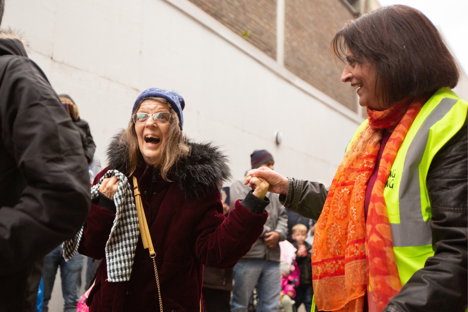 Two women share a moment of joy at an outdoor community event. One wears a high-visibility vest and bright scarf, holding hands with another woman who laughs warmly, dressed in a winter coat and hat.