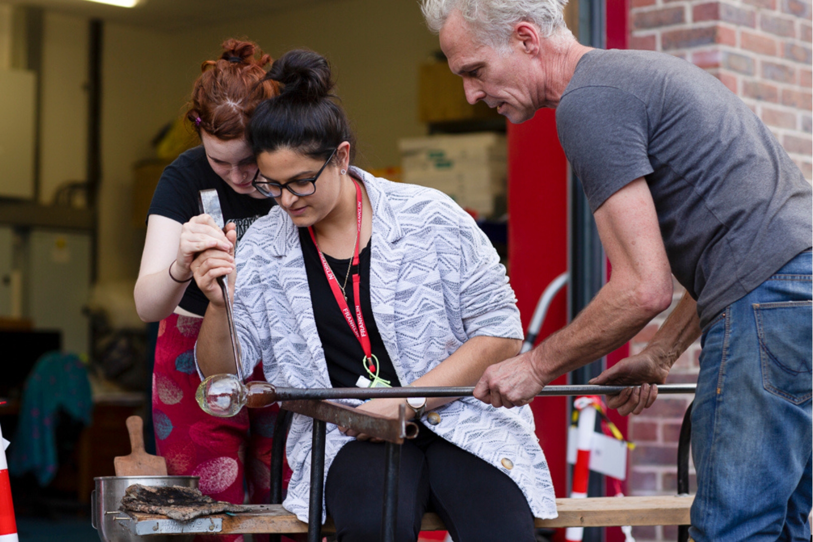 Three people collaborate on glassblowing. A woman seated at a bench shapes molten glass on a metal rod, guided by two instructors on either side in an open workshop space.